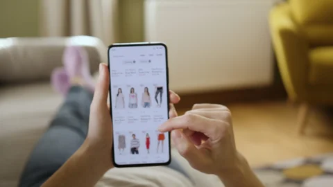A person relaxing on a sofa while browsing and shopping for clothes on a smartphone app.